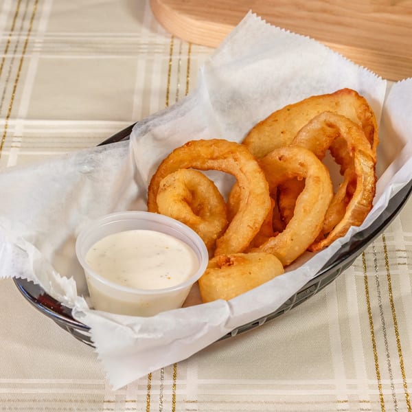 Golden-brown onion rings in a basket with a side of creamy dipping sauce on a checkered tablecloth.