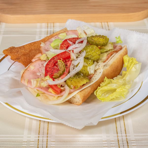 A Philly sub with ham, cheese, lettuce, tomatoes, onions, and pickles on a white plate.